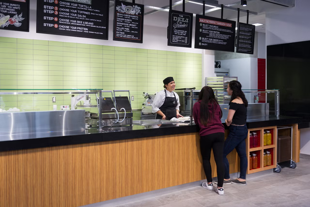 A Food Services worker behind a counter helps two customers.