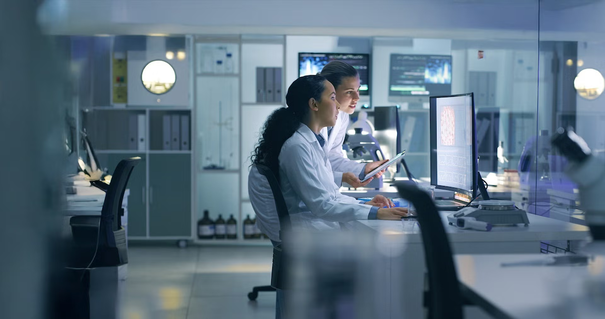 Two women in lab coats look at a computer monitor in a lab environment.