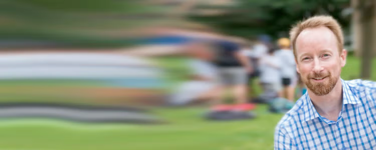 Dr. Troy Glover stands on a grassy field with distorted images of activity behind him.