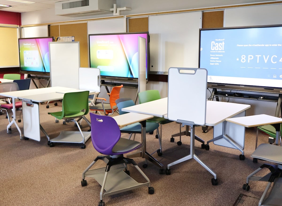 A modern classroom setup with multiple tables and chairs in various colours, two large screens displaying content, and a whiteboard.