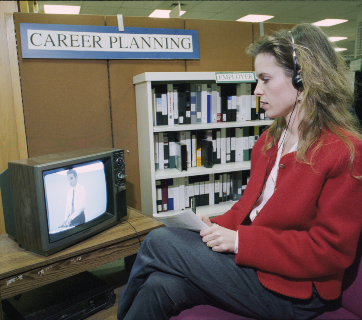 A young woman wearing headphones watches a video tape on a black and white TV with a "career planning" sign above.