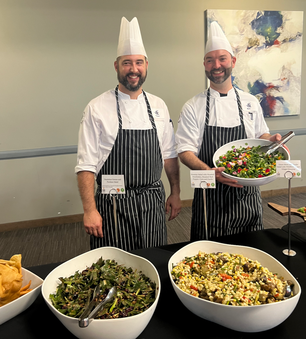 Two Catering chefs in white hats and striped aprons stand behind a table with large bowls of salad