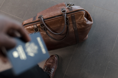 A person holds a passport with a duffel bag at their feet.