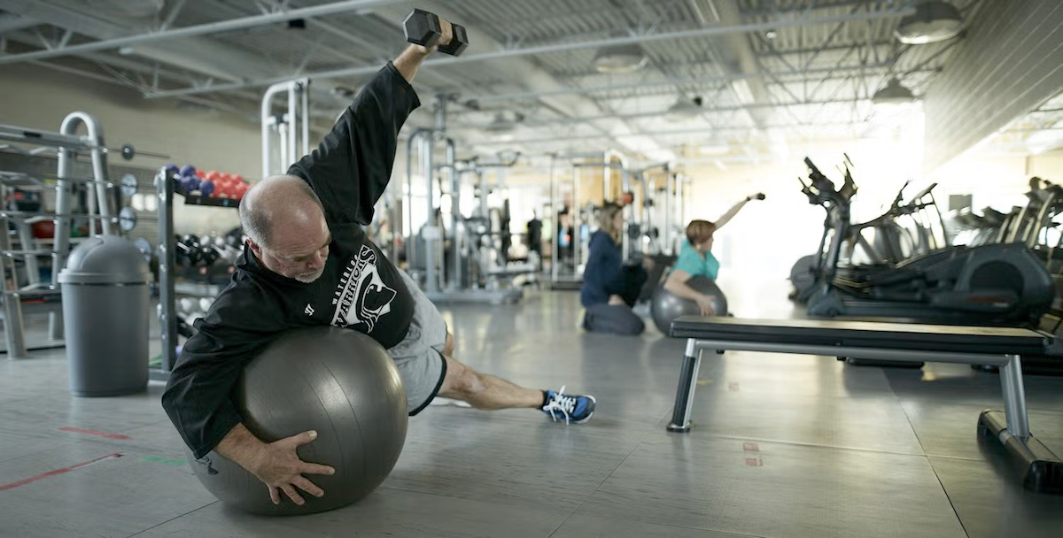 A man raises a dumbbell while planking on an exercise ball in a gym environment.