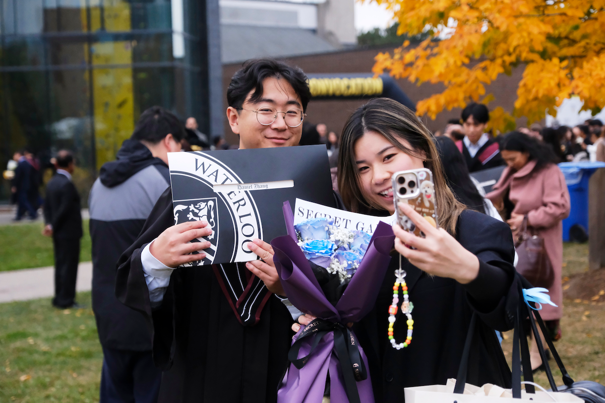 Two smiling grads in convocation regalia take a selfie after receiving their diplomas.