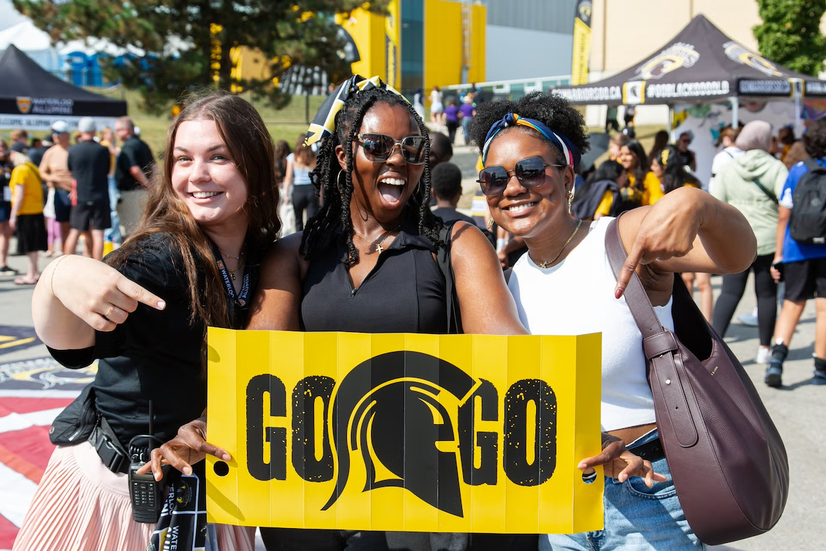 Three women hold up a "Go! Go!" sign with the Warriors logo.