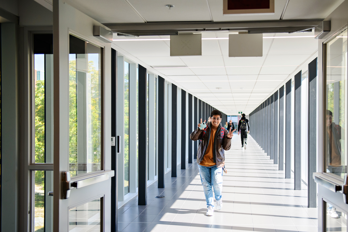 A student smiles and flashes peace signs as they walk on a campus overpass.
