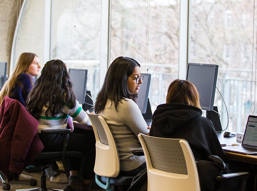 Four individuals are sitting at computer desks, facing away from the camera, in a well-lit space with large windows.