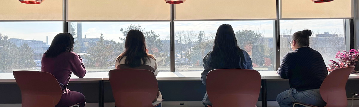 Four women sit at chairs against a counter with windows in front of them.