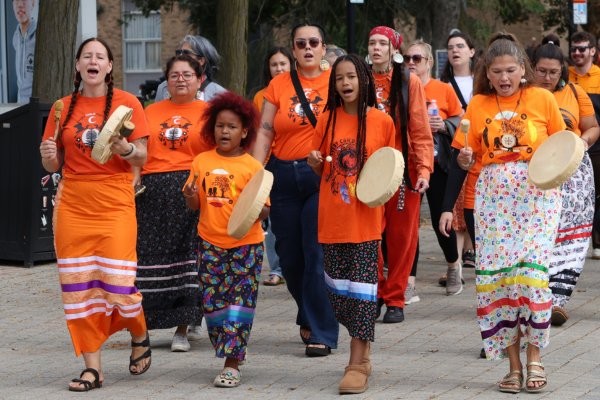 Participants wearing orange shirts walk together while playing hand drums during an Indigenous community gathering.
