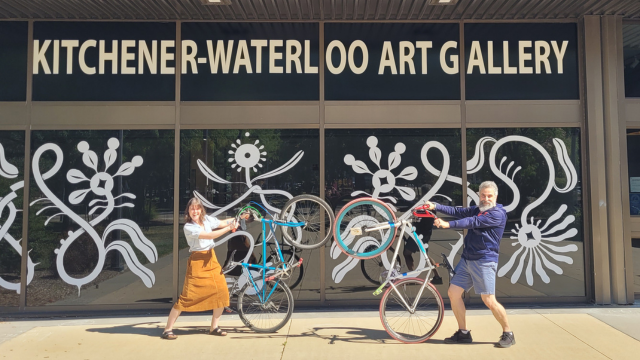 Two people pop wheeliers on their bikes outside the Kitchener Waterloo Art Gallery entrance.