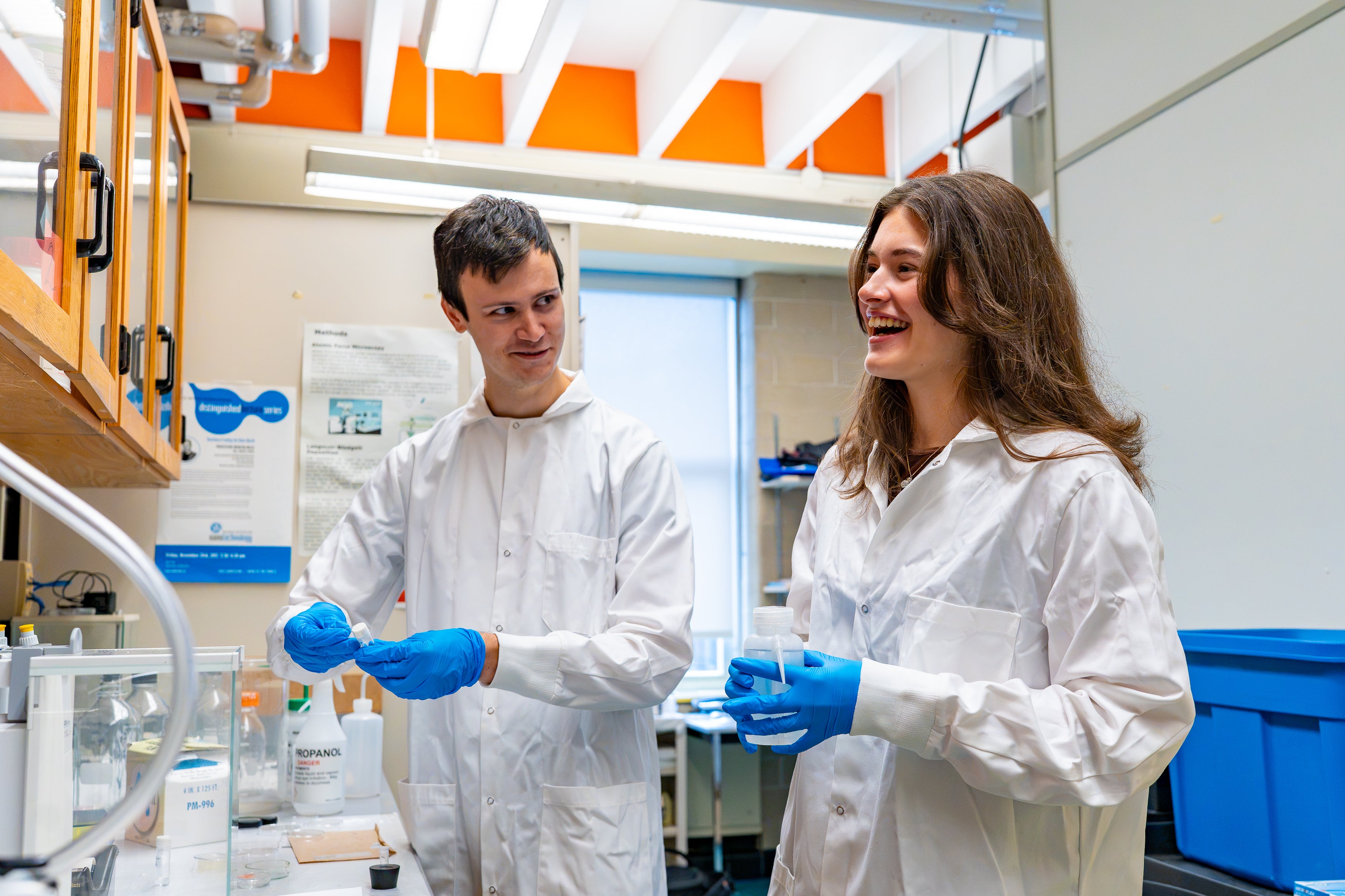 Two researchers wearing lab coats and blue gloves work together at a laboratory bench surrounded by scientific equipment and storage bins.