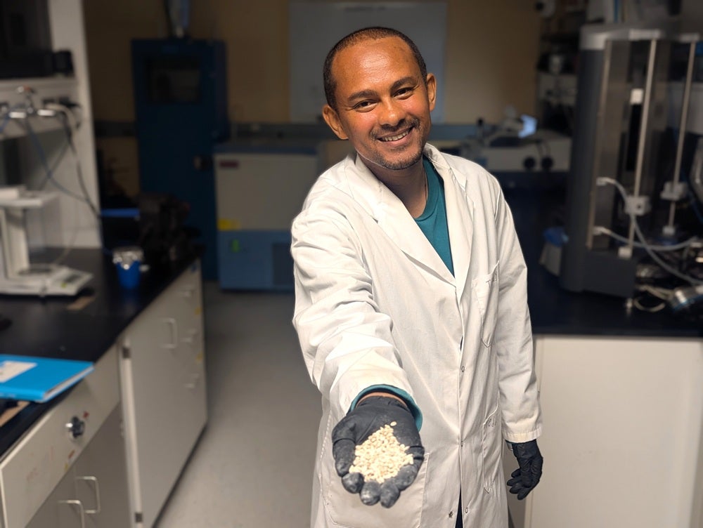 Researcher in a laboratory wearing a white lab coat and gloves holds a handful of pellet-like material while standing among scientific equipment.