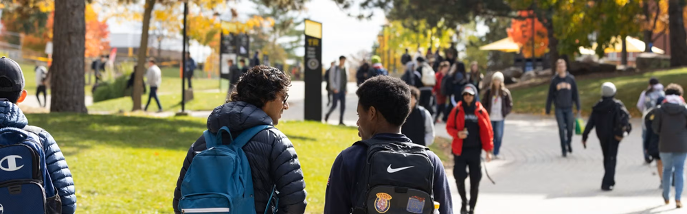 Students walk along a tree-lined campus pathway on a sunny autumn day, with colourful foliage and groups of people moving between classes.