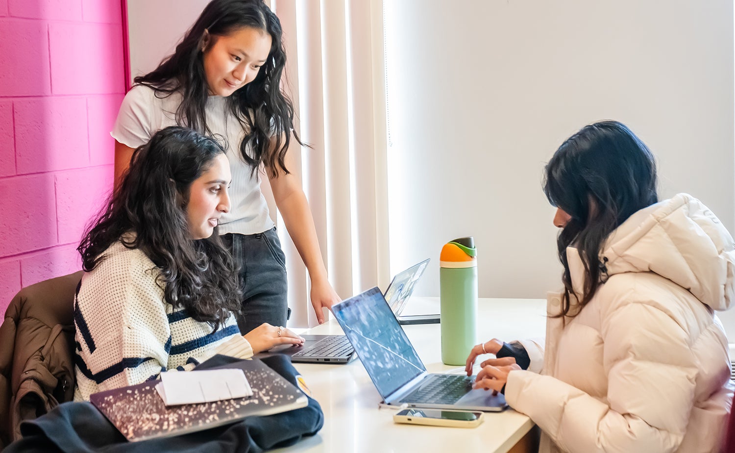 Three students work together on laptops at a table in a bright study space at Dana Porter Library.