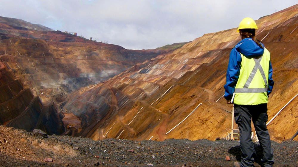 Person wearing a hard hat and high-visibility vest overlooks a large open-pit mining operation.