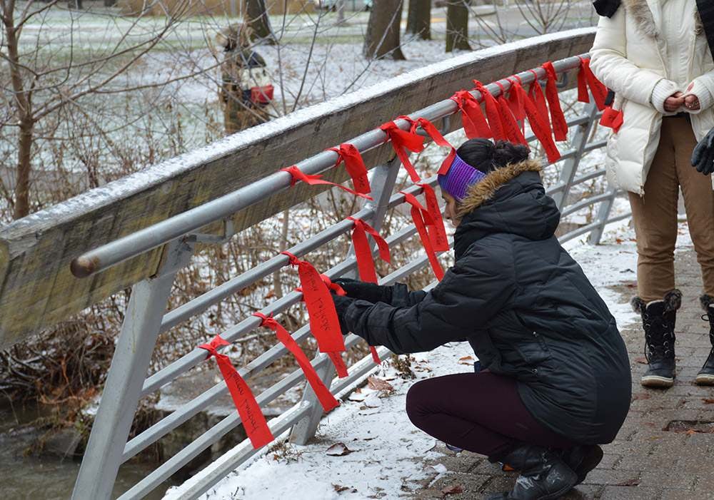 Person in a winter coat ties a red ribbon to a snow-dusted bridge railing alongside many other red ribbons.