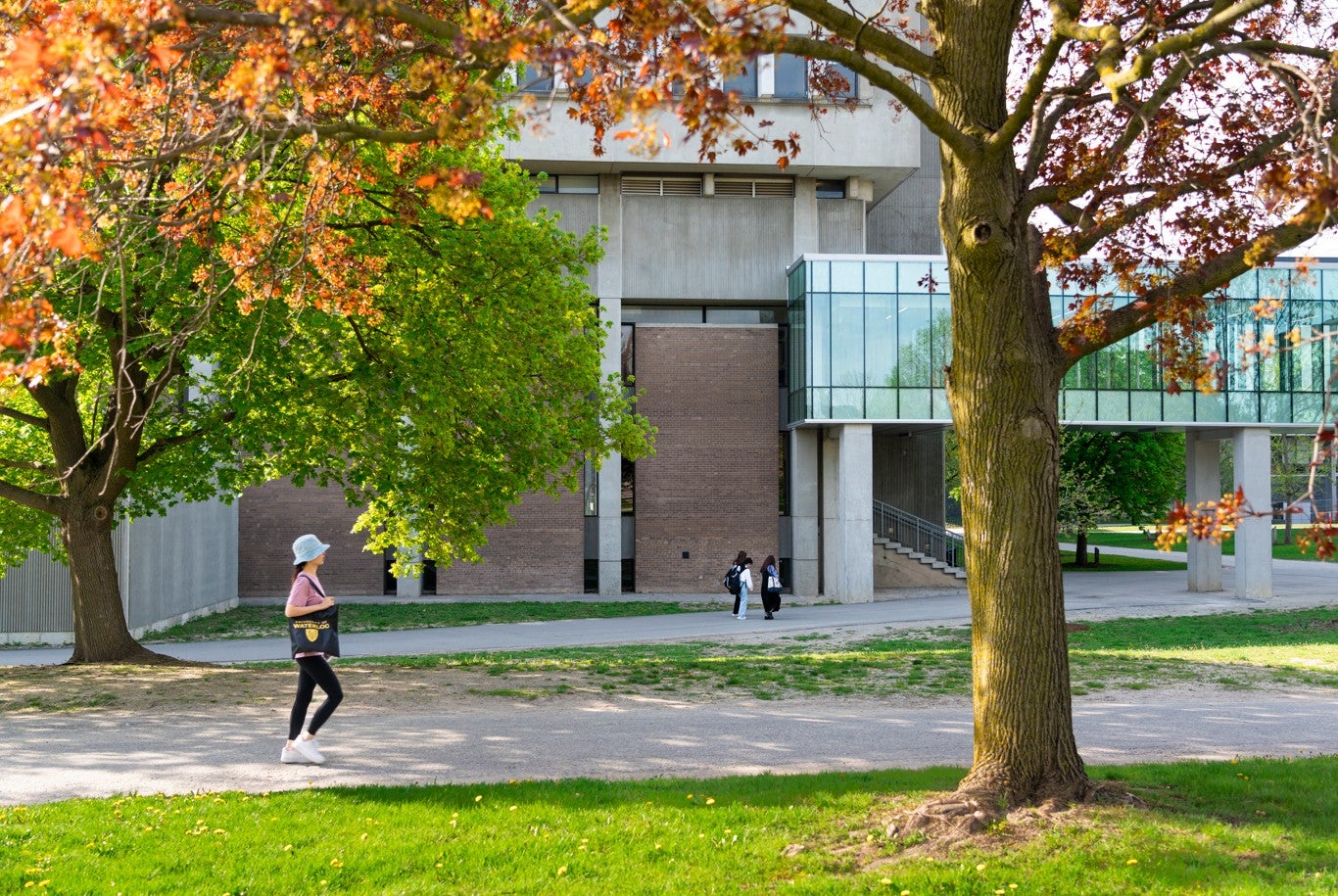 Copilot said: Student walks across a green campus lawn framed by leafy trees, with an academic building and pedestrian walkway in the background.