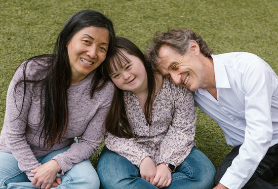 Persons seated together on a grassy lawn outdoors, posing closely for a group portrait in a park-like setting.
