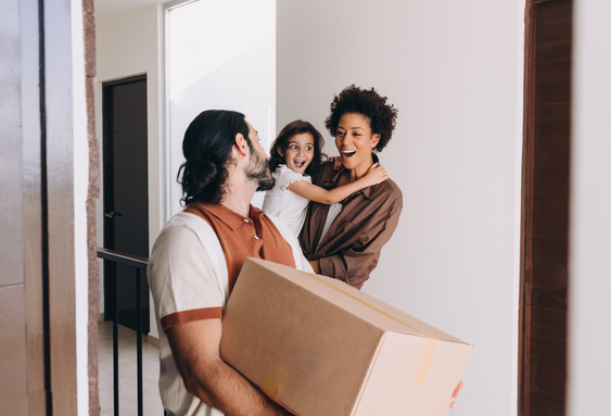 Person carrying a moving box enters a home while two other people stand nearby in an interior hallway.