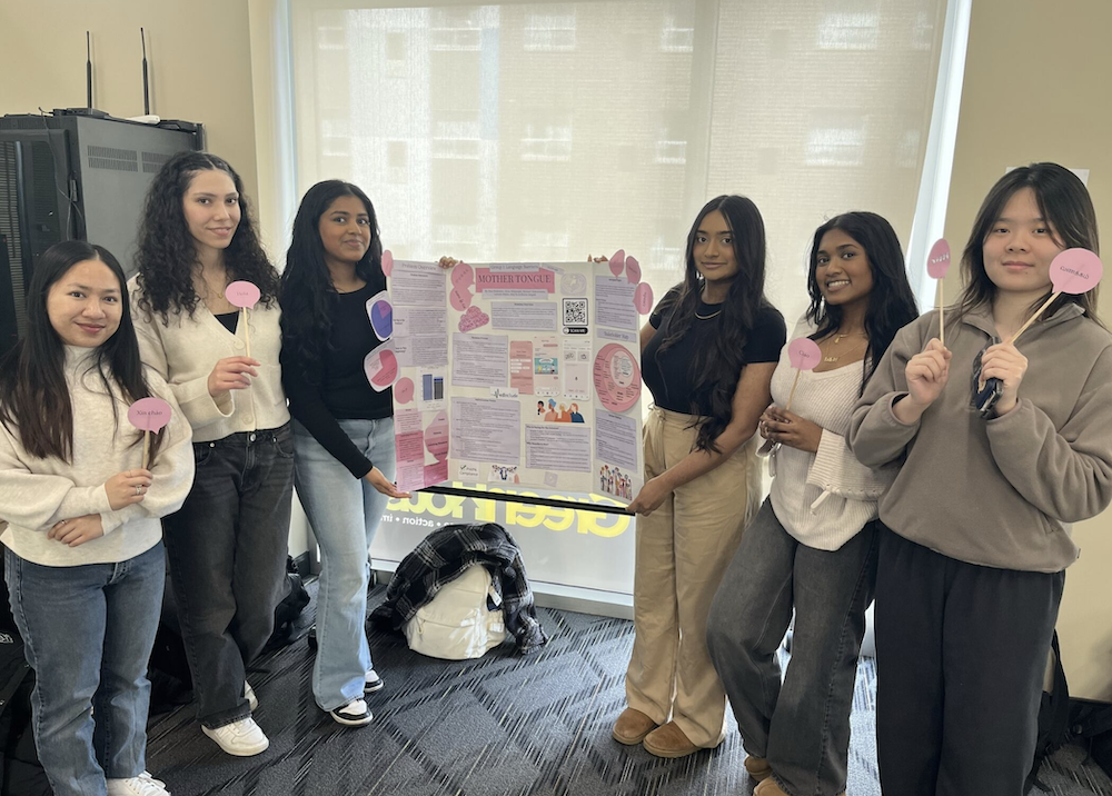 Six young women stand next to their poster display while holding cartoon hearts on sticks.