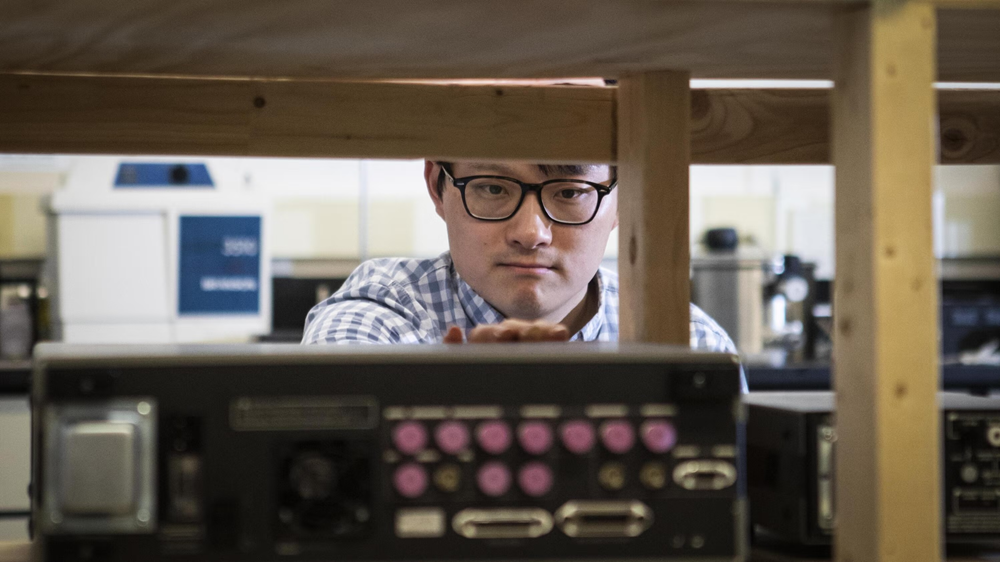 Person examines electronic equipment mounted beneath a wooden structure in a laboratory or research setting.