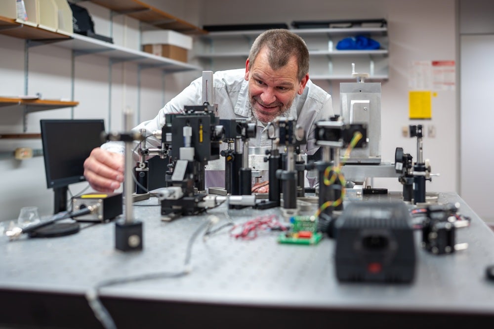 Person adjusts precision laboratory equipment on a research workbench surrounded by optical and electronic instruments in a university lab.