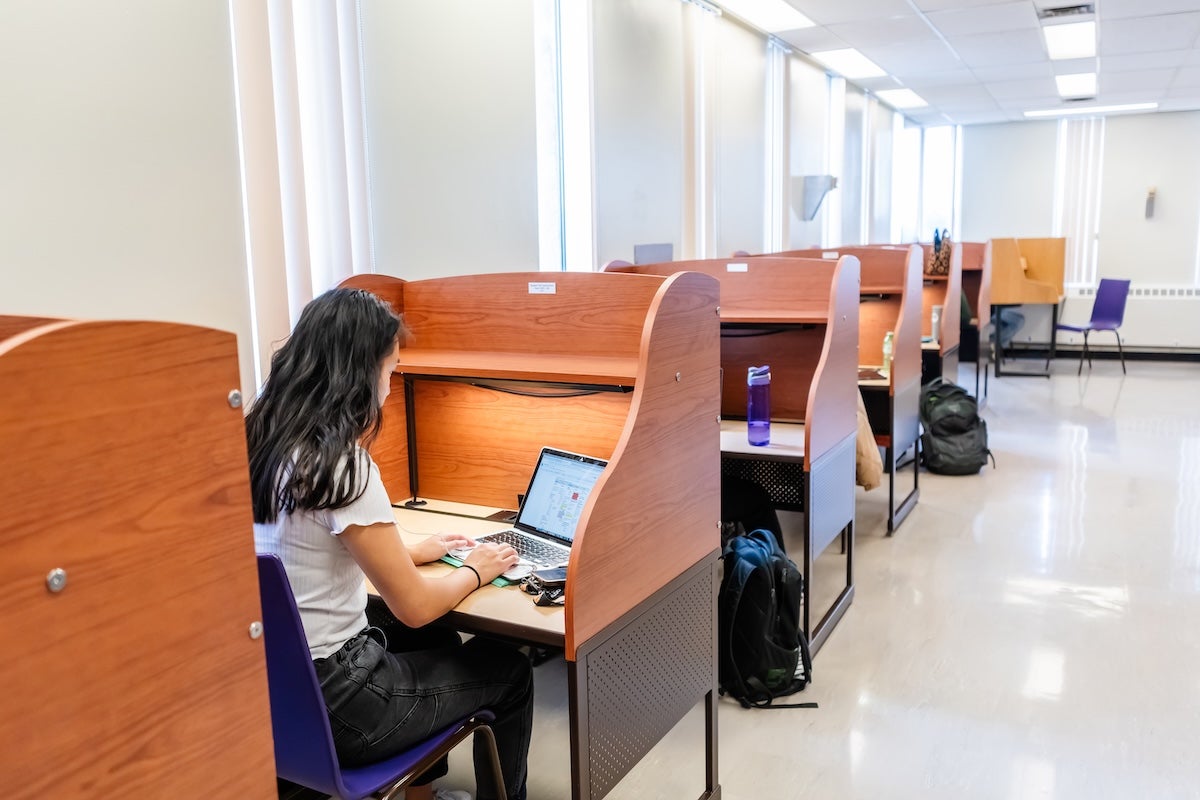 A student with long dark hair is sitting at a study desk using a laptop in a quiet, well-lit room.