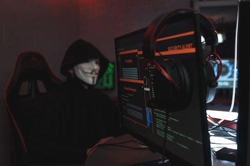 A hacker in a Guy Fawkes mask sits next to a bank of computer screens.