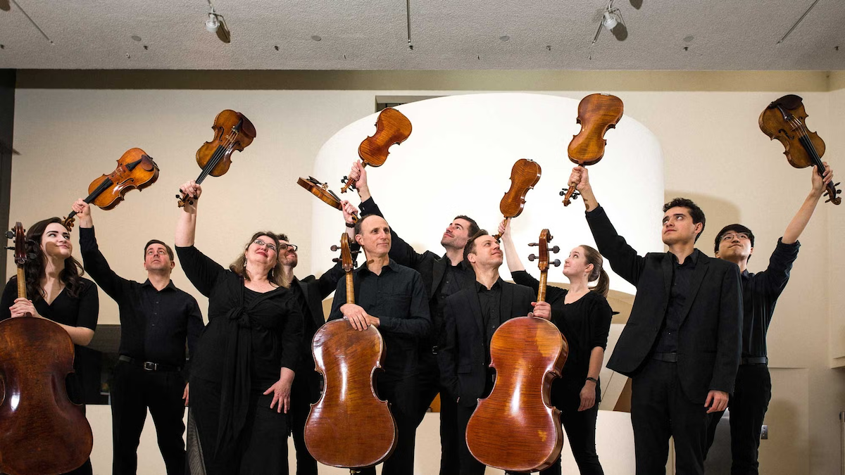 Members of the Magisterra Ensemble hold their stringed instruments, with some performers holding violins overhead.