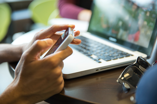 A person uses a smartphone with a laptop in the background.