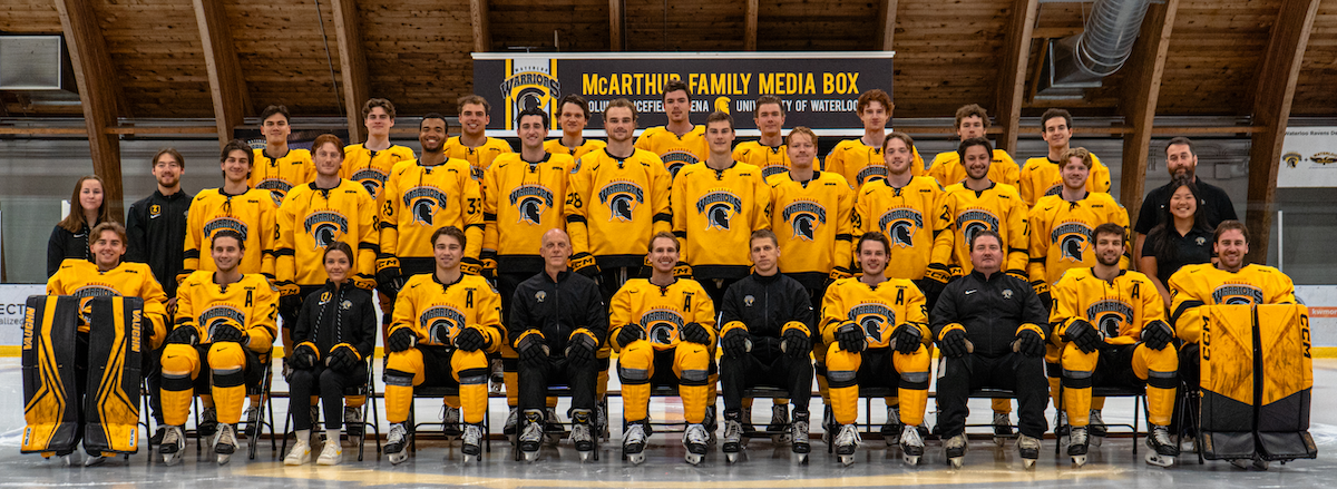 The men's hockey team stands in front of the McArthur Family Press Box in the CIF Arena.