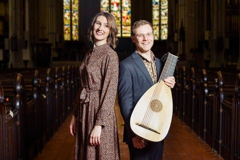 Sinéad White and Jonathan Stuchbery stand in the aisle of a church. Jonathan holds a lute.