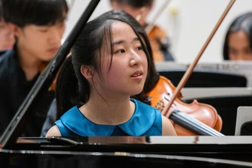 Catherine He sits at the piano with an orchestra behind her.