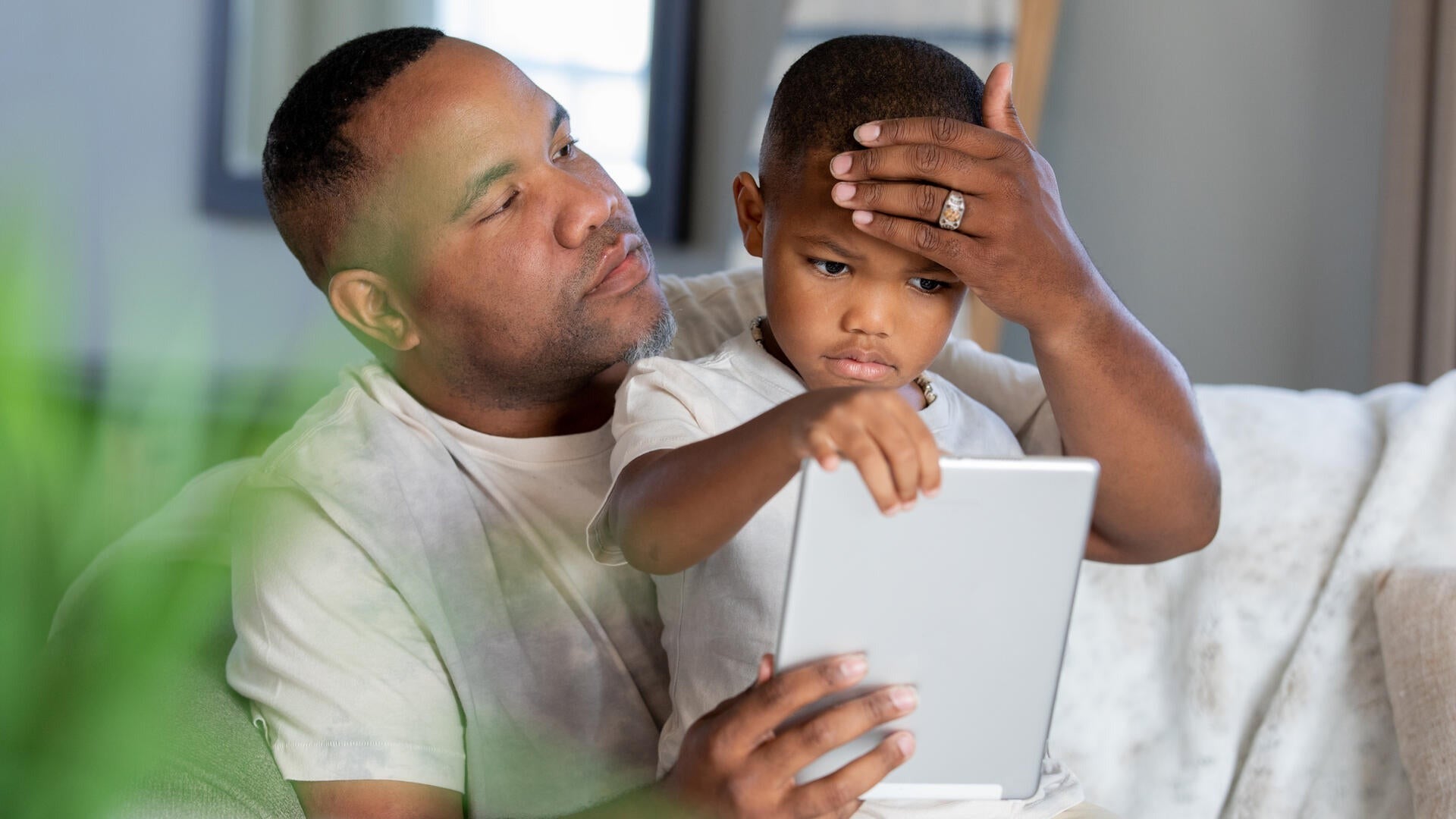 A father checks his son's temperature while the boy looks at a tablet.
