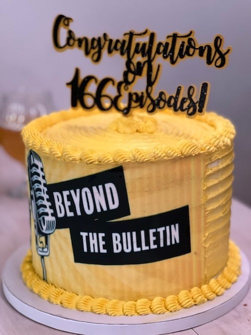 A tall cake with yellow frosting with the Beyond the Bulletin logo silkscreened on the side and a topper that says &quot;Congratulations on 166 episodes!&quot;