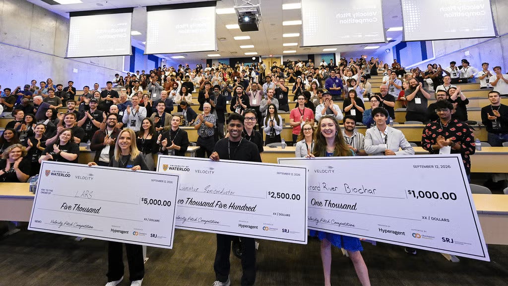 Three students stand at the front of a class with big cheques