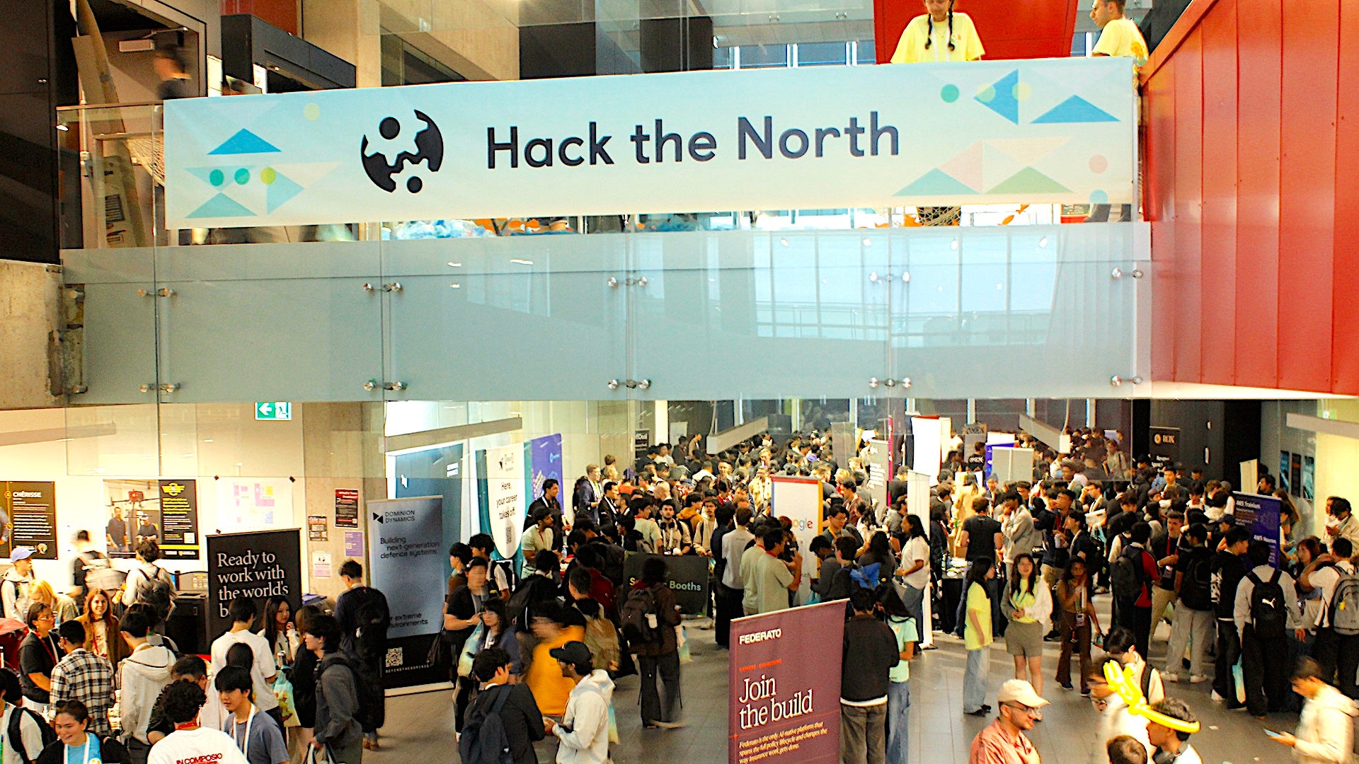 A large crowd of students gathers beneath a prominent “Hack the North” banner, with booths, displays, and networking activity filling the space.