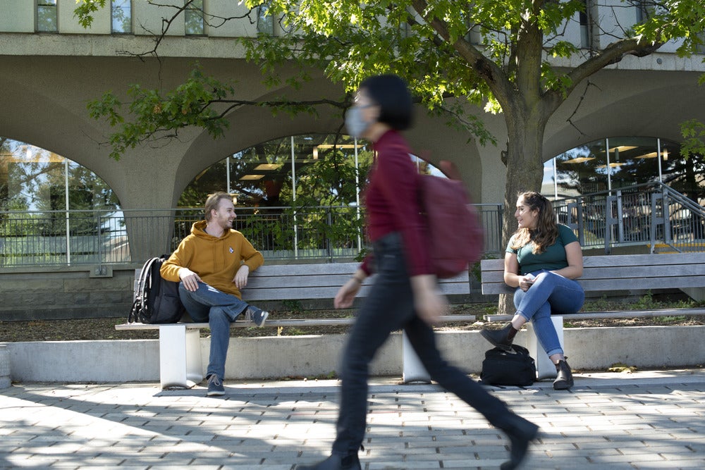 Students sit on a bench in front of the Dana Porter library. A slightly blurred student can be seen walking past the bench in the foreground.