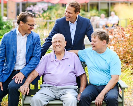 Dr. Ron Schlegel sits on a bench outdoors with his three sons gathered around him in a garden setting.