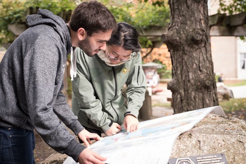Two students look at a map unfolded over a rock in a forest. 
