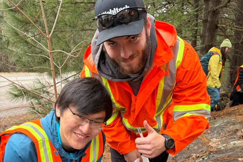 Two male students crouching next to a rock formation, one is giving a thumbs up. 