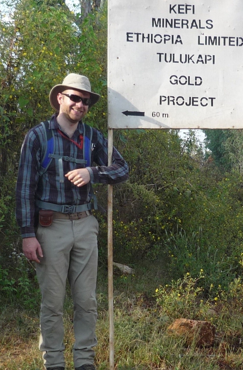 Chris is standing on a trail surrounded by greenery. He is wearing a hat, sunglasses, a plaid shirt, hiking pants and boots and is carrying a backpack. His arm is looped around a sign that says &quot;Kefi Minerals Ethiopia Limited Tulukapi Gold Project."
