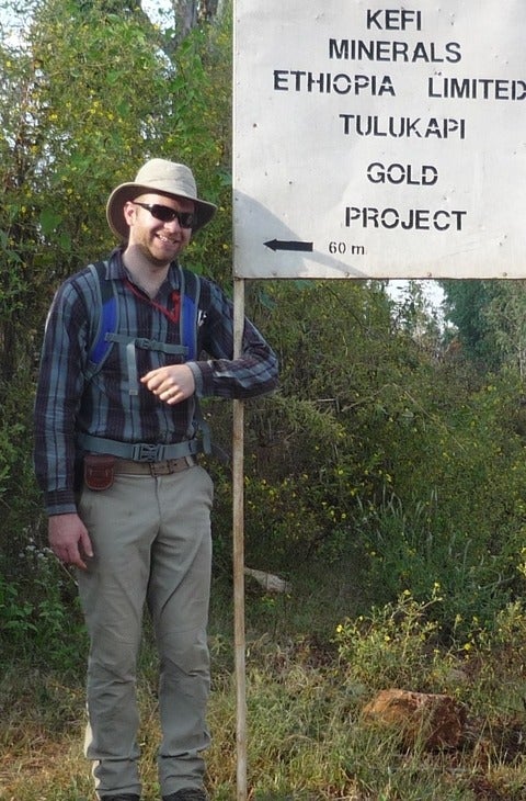 Chris is standing on a trail surrounded by greenery. He is wearing a hat, sunglasses, a plaid shirt, hiking pants and boots and is carrying a backpack. His arm is looped around a sign that says &quot;Kefi Minerals Ethiopia Limited Tulukapi Gold Project."