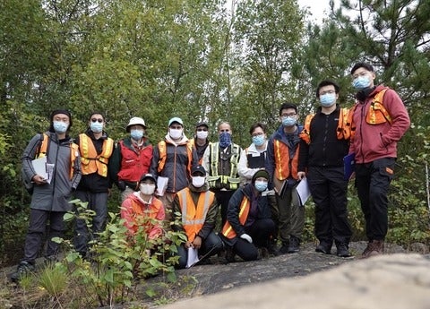 Group of students, TAs, and professor standing on a rock surrounded by trees. All are wearing masks