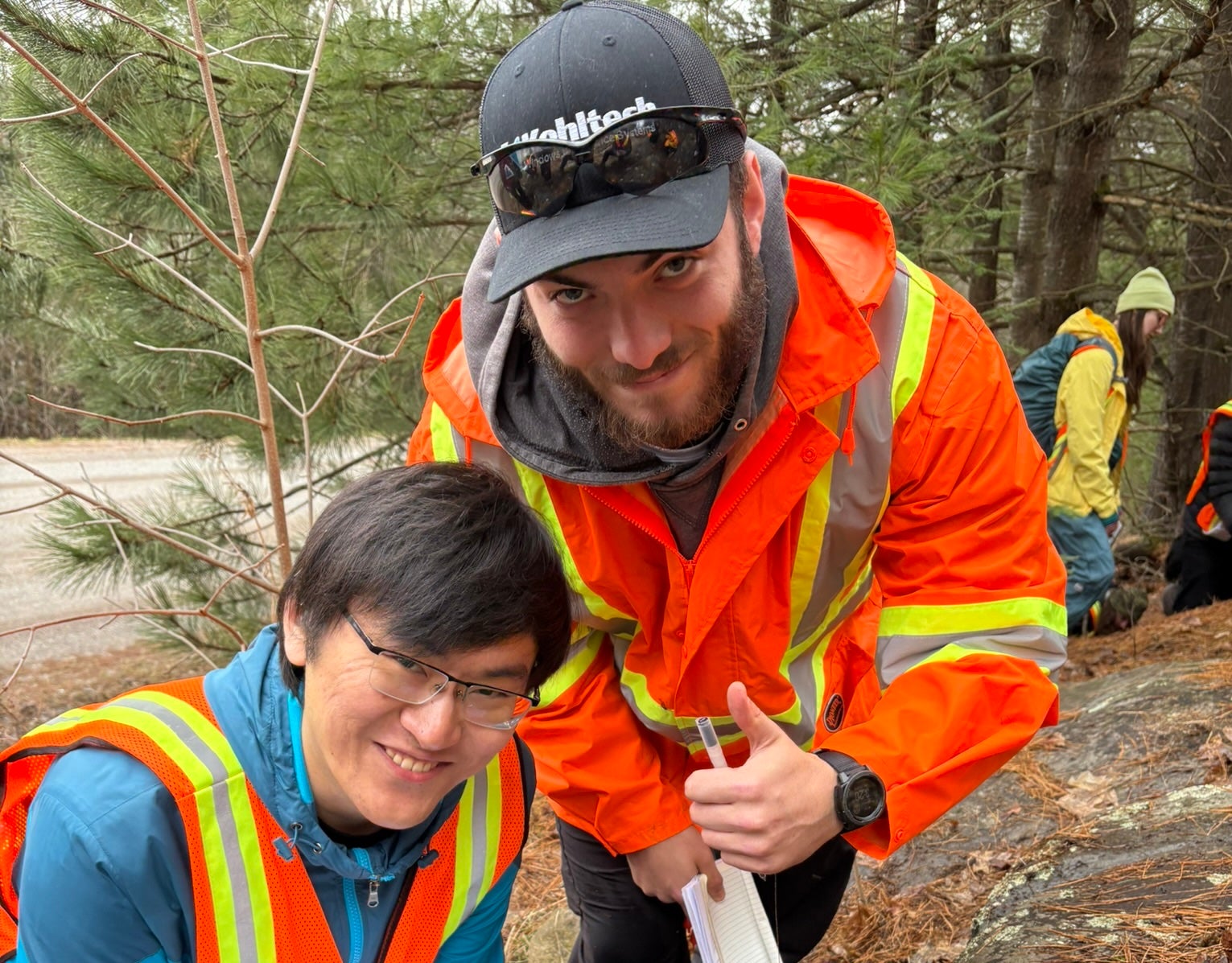 Two male students crouching next to a rock formation, one is giving a thumbs up. 