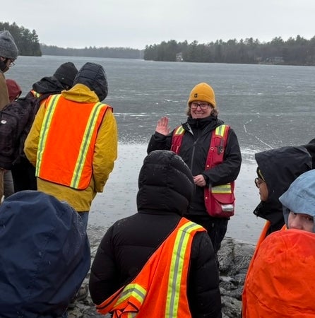 Instructor Jen Parks waves from in front of a group of students on the edge of a body of water. 