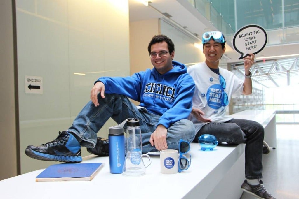 Manuel and Ryan sitting on a white desk in University of Waterloo Science swag