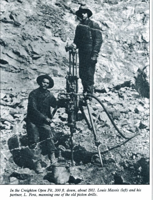 two men operating a piston drill on the surface of an open pit that is 300 feet down