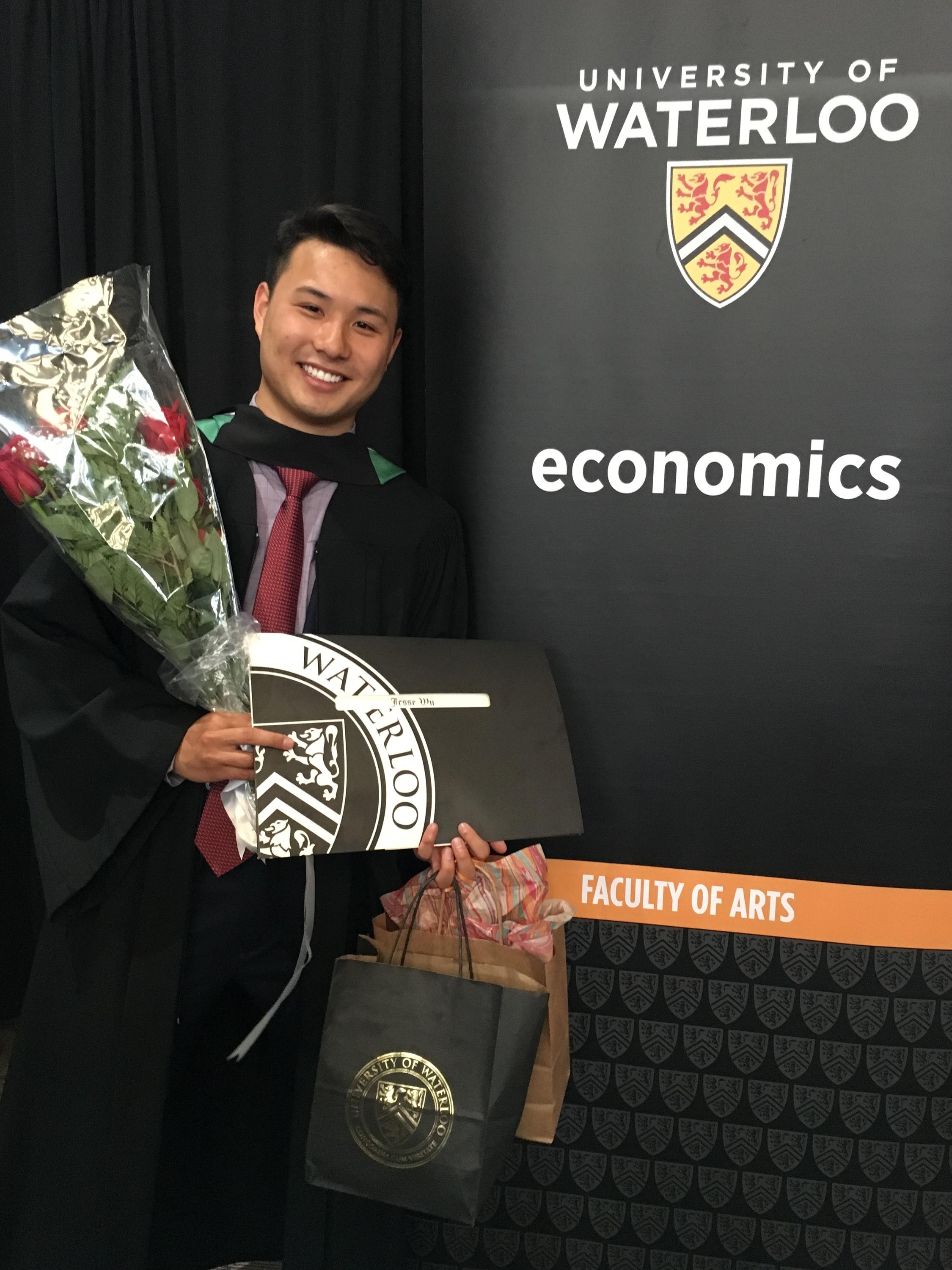 Student holding diploma and flowers in front of sign that says, "Economics"